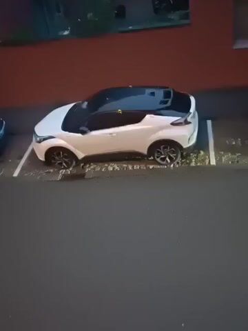 A white Toyota C-HR parked in a parking spot. The car has a black roof and tinted windows. A black SUV drives past in the background.