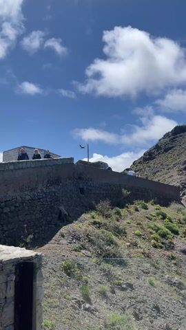 The video shows a large bird, possibly an eagle or vulture, flying in front of a stone wall and a steep, rocky hillside. Several people stand on the wall looking at the bird. Cars are parked behind them. The bird flies to the right of the frame, descending towards the hill. The sky is blue with scattered white clouds. Later, the camera focuses on a man in a denim jacket and cap, his back to the camera, observing the surroundings. The setting appears to be a mountainous region on a sunny day.