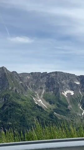 The video shows a view of a mountain range with green slopes and rocky peaks. Patches of snow are visible on the mountainside. In the foreground, a metal guardrail and a striped barrier pole are present, suggesting the perspective is from a road or viewpoint. The sky is cloudy with some blue patches. The camera appears to be moving, as indicated by slight shifts in the scene and the presence of the barrier pole moving across the frame. The overall environment is mountainous and natural, likely in a high-altitude region.