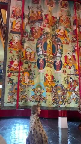 This video shows a wide shot of a room with a large, colorful tapestry hanging on the wall. The tapestry depicts a Buddhist mandala with many figures. A person wearing a patterned robe is kneeling in front of the tapestry, looking up at it. The room appears to be a temple or monastery, with ornate decorations and murals on the walls. The floor is tiled and reflective, and there are wooden beams and columns supporting the ceiling. The lighting suggests it's daytime, possibly late afternoon, as there are warm tones in the light. The atmosphere seems peaceful and contemplative, with the person engrossed in observing the tapestry. The setting strongly suggests a religious or cultural site in India, aligning with the provided location context of Fatehpur. There is no visible text in the video.