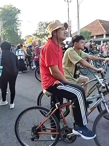The video captures a lively street scene, bustling with a large crowd of people, some walking, some on bicycles. The camera follows individuals and groups as they move through the area, which appears to be a public space or market due to the presence of vendors and stalls. One man in a red shirt and a bucket hat is prominently featured, riding a bicycle alongside a younger boy on another bike. The sun is bright, creating lens flares at various points in the footage. The surroundings include buildings, trees, and streetlights, indicating an urban or semi-urban setting. The atmosphere suggests a community event or a busy day out.
