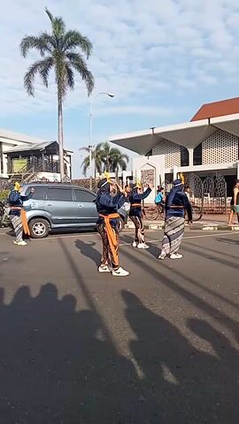 The video depicts a vertical street scene on a sunny day with a few clouds. Four men dressed in traditional clothing stand and walk along the side of the road, seemingly performing or participating in an event. One man stands next to a parked grey van. In the background, a building that looks like a mosque is visible. A palm tree stands beside the road. In the distance, a few people are on bicycles. Near the mosque, a puff of smoke is visible, possibly from a pyrotechnic device.