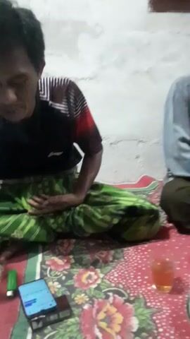 The video shows a gathering of several men sitting on the floor, likely in a home in Panjalin Kidul, Indonesia. The room appears to be simple, with a concrete wall and a patterned rug covering the floor. The men are dressed in casual clothing, with some wearing traditional Islamic caps.

On the rug in front of the men are several plates of food, including what appears to be fruit, fried snacks, and possibly some savory dishes. There are also glasses of a reddish liquid, possibly tea or juice, and a pack of tissues. A mobile phone and a lighter are visible on a smaller rug in the foreground.

The men are engaged in conversation, though their exact topic is unclear. Some appear to be listening attentively, while others are speaking. The atmosphere seems relaxed and informal, suggesting a social gathering or a meal shared among friends or family. The lighting suggests it could be daytime or early evening.

There is no visible text in the video.