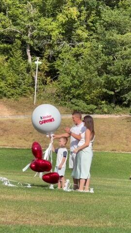 A family of three, a man, a woman, and a young boy, stand on a grassy field with trees in the background. They are holding onto a large white balloon with the text "Kinder Opmr3" printed on it in red. Below the large balloon, there are several red heart-shaped balloons. As the man and woman pull on the string of the large balloon, it bursts, releasing a cloud of white confetti and smaller pink and white balloons into the air.