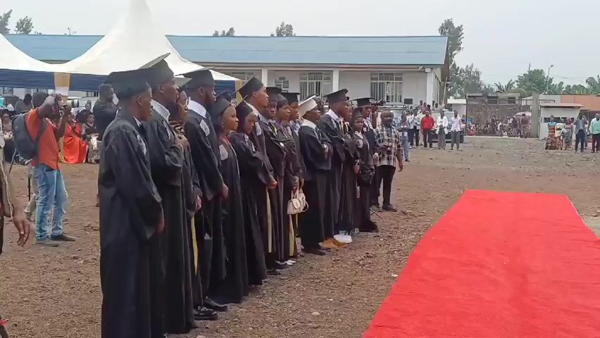 A group of people wearing graduation robes and caps are standing in a line in front of a red carpet. They are facing forward. To the left of the group, a man is filming them with a video camera on a tripod. Other people are standing around watching. Behind the group is a white tent and a building with a blue roof. The sky is overcast.