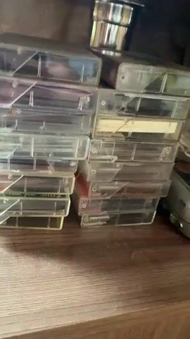 The video shows a stack of cassette tapes sitting on a wooden shelf, seemingly inside a cabinet or beneath something. The cassettes are mostly in clear plastic cases, and the spines of the cases have various colorful labels indicating the albums or artists on the tapes. Some of the visible names include Николай Басков (Nikolai Baskov), Союз (Soyuz), and Любимой (Favorite). There is also a cylindrical, metallic object visible in the top-left of the frame, possibly a part of a furniture leg or fixture. The audio contains indistinct speech.