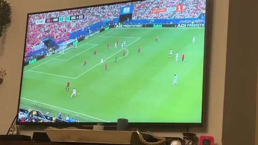 A television screen displays a soccer match in progress. The scoreboard at the top left indicates the game is at 17:38 with the score NOR 0 ENG 0. The field is green, with white lines marking the boundaries and penalty areas. Players in red and white uniforms are visible on the field. The crowd in the background is wearing red. The screen is mounted on a wall above a fireplace mantel. On the mantel, there are decorative items, including a woven tray, a candle, and small figurines.