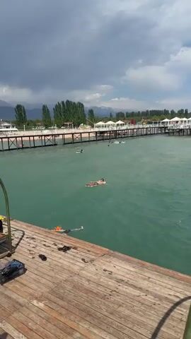 The video opens with a medium shot of a wooden pier with two people swimming in the clear, turquoise water. The pier is lined with white cabanas and a long wooden walkway stretches into the distance, with more people swimming and some on inflatable tubes. In the background, trees and mountains are visible under a cloudy sky. The camera then pans to a wider shot of the lake, showing more of the pier and the surrounding landscape. The water is calm, with ripples from the swimmers. The video then cuts to a shot of a clear blue sky and the ocean. A metal railing is visible in the foreground. The water is a deep blue-green, and there are whitecaps on the waves. In the distance, a small boat can be seen.