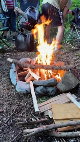 A campfire is burning in a forest clearing. The fire is built within a ring of stones and surrounded by wood and kindling. In the background, several people are visible. One person, wearing black shorts and a light-colored t-shirt, is tending to the fire. Other individuals are partially visible, some sitting on camping chairs. Bags and other camping gear are also present in the scene. The ground is covered in leaves, twigs, and dirt.