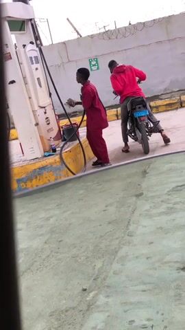 A male gas station attendant in a maroon uniform is holding a fuel nozzle attached to a pump. A male on a motorcycle, wearing a red hooded sweatshirt and a black baseball cap, is parked next to the attendant. The attendant appears to be handing something, possibly money, to the person on the motorcycle. The scene takes place at a petrol station with a concrete ground and a wall in the background.