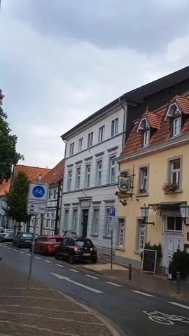 A street-level, panning video shows the exterior of a yellow building with a red-tiled roof. The building appears to be a hotel and restaurant named "Pilgrim Haus" with the founding date "seit 1904". A decorative figure of a soldier holding a rifle stands mounted on the side of the building. Another building to the left is white with a darker roof. Flower boxes with pink flowers are attached to the windows of the yellow building. A street with white lines and a bike lane is visible in the foreground. The sky is overcast.