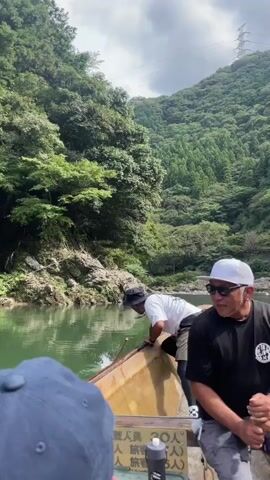 Two men are in a boat on a body of water. The man in the foreground is wearing a black t-shirt, a white hat, and sunglasses. He is holding a paddle. The man in the background is wearing a white t-shirt, a bucket hat, and shorts. He is holding a long pole and appears to be interacting with the water. The boat has Japanese writing on it. The background consists of lush green hills and trees, with a cloudy sky above. The water is green and calm.