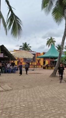 The video captures an outdoor gathering at what appears to be Pulodolang, Indonesia. The scene is set on a sandy ground with a patterned paved area in the foreground. A stage with an elaborate backdrop featuring yellow and orange motifs and what looks like Arabic calligraphy is visible in the center, covered by a large, light-colored tent or umbrella. To the right of the stage, a teal-colored tent provides further shelter.

Several people are present, mostly men and boys, dressed in traditional attire, including sarongs. They are seen walking around the area and congregating. Some are standing near blue plastic chairs, suggesting a seating arrangement for an event. Coconut palm trees are prominent in the frame, indicating a tropical location. In the background, there are more tents, trees, and what looks like a communication tower. A motorcycle is parked to the right of the teal tent.

The sky is overcast, suggesting it might be daytime but not necessarily bright sunlight. The overall atmosphere seems relaxed, with people moving about and engaging in conversation or preparation for an event. The image is likely taken during the day, given the ambient light. There are no overt expressions of strong emotion, but the gathering suggests a community event or ceremony. There are no clear signs of distress or urgency.