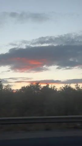 This is a video recording of a sunset. The sky is filled with clouds, some of which are illuminated with hues of orange and pink. Below the sky, a silhouette of trees is visible against the horizon. In the foreground, a dark railing is present. The camera appears to be moving, possibly from a vehicle.