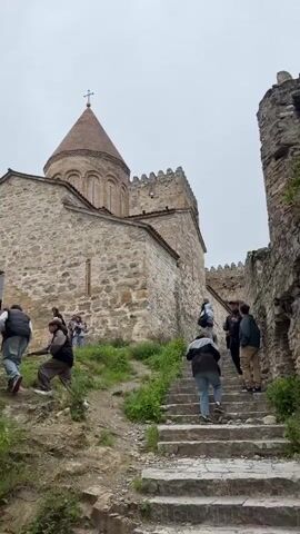 The video shows a group of people visiting a stone church on a hill. Some people are walking up the stone stairs, while others are standing near the church taking pictures. The church has a conical roof with a cross on top. The sky is overcast. The video is shot from a low angle, looking up at the church and the people. There is a comment on the video about someone named “Zhenya” believing in everything.