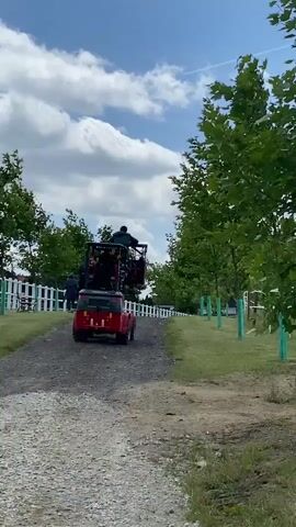 A red utility vehicle with a person in the driver's seat and another person in a raised cage-like structure behind them is driving away from the camera down a gravel path. To the left of the path is a white picket fence, and beyond that, some trees and a small white building. To the right of the path is a grassy area with trees. The sky is blue with white clouds.