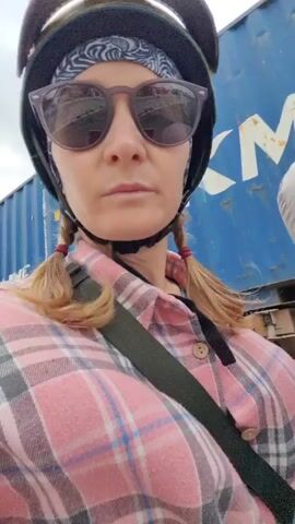 The video shows a woman riding on the back of a motorbike. She is wearing a yellow helmet, sunglasses, a bandana, and a pink plaid shirt. She has pigtails. The motorbike is driving alongside a large white semi-truck and a blue shipping container. The driver of the motorbike is wearing a light grey jacket and a purple helmet. The sky is overcast.