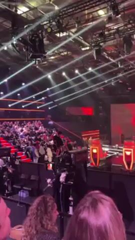 The scene captures an entertainment event in a large venue filled with an audience seated on tiered rows of red chairs. Above the crowd are bright spotlights that create a network of sharp lines across the ceiling. A person in a dark outfit uses a zip line to descend from a higher level toward the stage area, where iconic red chairs are positioned. Several individuals stand near the stage. Multiple cameras and other technical equipment are positioned around the stage area to film the event. The audience appears engaged, though details of their expressions and interactions are limited due to distance and resolution.