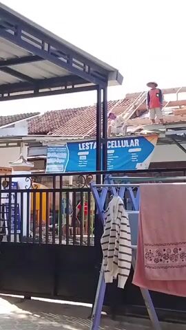 The video shows a group of construction workers on the roof of a building. The building appears to be undergoing some kind of renovation or repair, as some of the roof tiles are missing and the wooden frame is exposed. There are at least four workers visible, wearing casual clothing and hats. One worker is standing on the roof, while two others are actively working on the tiles. Another worker is sitting down, possibly taking a break. In the foreground, there is a clothesline with several towels hanging on it, partially obscuring the view of the construction. A sign that says "LESTA CELLULAR" is visible on the building behind the workers. The overall environment appears to be an outdoor setting on a bright, sunny day.