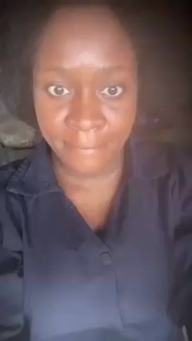 A medium shot of a dark-skinned woman wearing a dark navy button-down shirt. She looks directly into the camera. At first, her expression is neutral, then she rolls her eyes down and gives a subtle smile. Behind her is a dark setting, with a pillow visible on a surface. The video has a slightly grainy quality.