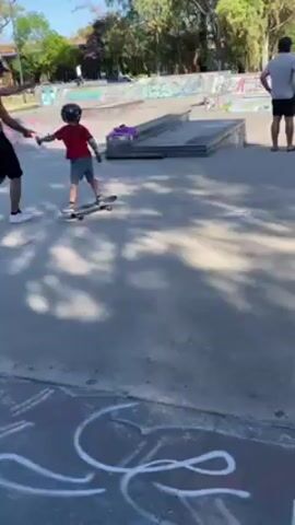 The video shows a young child skateboarding at a skate park. The child is wearing a red shirt, dark shorts, a helmet, and elbow pads. There is a man next to the child, apparently giving instructions, and two other people are standing nearby watching. The skate park has various ramps, rails, and graffiti-covered surfaces. It is a sunny day, and there are trees visible in the background. The child is riding on a skateboard across a flat concrete area.