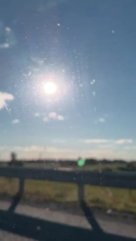 A person is speaking in Russian from inside a vehicle, with the sun shining brightly in a clear blue sky visible through the windshield. The windshield has some smudges and streaks. In the background, a blurred landscape with trees and a fence can be seen, indicating the vehicle is in motion.