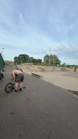 The video shows a skate park with several ramps and rails. At first, a shirtless man is bent over a bicycle, apparently making repairs. In the background, several people are using the park. The camera then shifts to a group of people sitting on the pavement. A young man with long curly hair, wearing a soccer jersey, is shown rolling what appears to be a joint. Other members of the group look on. Some of the group are sitting on scooters.