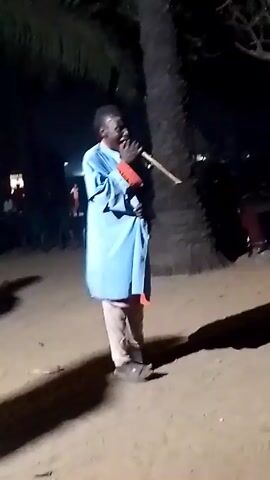 The video shows a person dressed in a long, light blue garment with red cuffs, light-colored pants, and dark shoes, standing in a sandy area at night. The person is playing a long, thin instrument, possibly a flute or pipe, held to their mouth. To the left of the person, there is a dimly lit building and a group of people in the background. To the right, a tall, dark palm tree stands behind the person, casting a shadow on the ground. The overall lighting is low, and the scene appears to be outdoors.