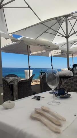 The video shows a sunny outdoor restaurant with a view of the ocean. There are several tables with white tablecloths and wicker chairs, shaded by large white umbrellas. Some of the umbrellas are branded with “Laurent-Perrier” and “TAITTINGER.” A waiter wearing a white shirt, apron, and blue vest walks in front of the camera. The ocean is calm and blue, and there is an island in the distance. On the table closest to the camera, there is a wine glass, keys, a small stone, and wrapped wooden utensils. People can be seen seated at other tables.