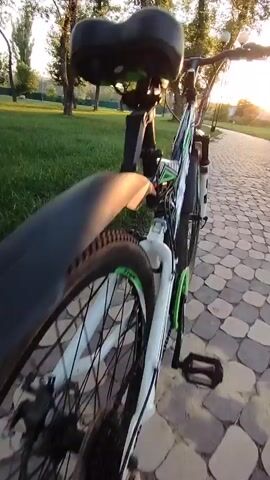 The video begins with a close-up shot of a bicycle's saddle and frame, then pans to show the handlebars and front wheel of a mountain bike. The bike is on a paved path lined with grass and trees. The sun is setting, casting a warm glow through the leaves. The camera then moves to a first-person perspective, showing the handlebars and a gear shifter. The bike is in motion, moving along the path. The video continues with shots of the bike from the same first-person perspective as it rides through a park-like setting. The sun is visible in the distance, creating a silhouette of the trees. The paving stones of the path are visible, as are the green blades of grass and the trunks of the trees. The overall impression is one of a peaceful and scenic bike ride at sunset.