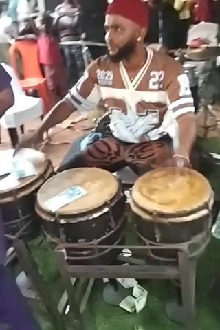 The video shows a live band performing at an outdoor event. The band consists of a drummer playing three conga drums with money placed on top of them, and a trumpeter. The drummer is wearing a jersey with the number 22 and the year 2025 on it. He also has a red hat. The trumpeter is wearing a black and white striped shirt. People are standing around watching the performance. There are white and orange chairs visible in the background. The ground is covered with grass and sand.