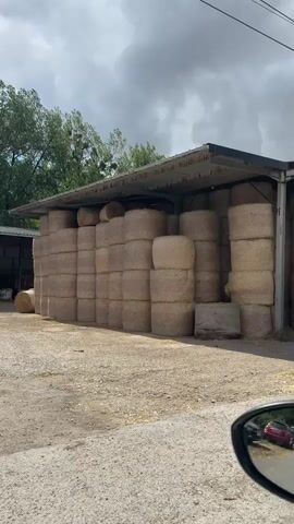 A camera pans across a rural landscape, focusing on a large stack of hay bales stored under a shed. The shed has a metal roof supported by wooden beams, and the hay bales are neatly stacked in rows, filling most of the space. In the background, a second, smaller shed is visible, containing some farm equipment. Trees with green foliage line the perimeter of the scene, and the sky is partly cloudy. The ground is covered in gravel and scattered hay. A small portion of a car's side mirror is visible in the foreground, suggesting the video was filmed from inside a vehicle.