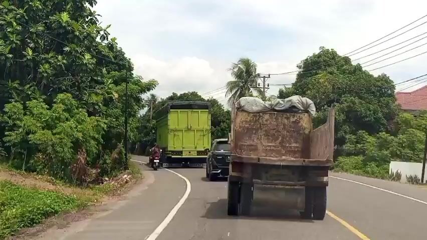 The video shows a view from a vehicle driving on a paved road. In front of the vehicle are two trucks. The truck closest to the camera is an older, rusty dump truck. Ahead of it is a lime green dump truck. To the left of the road, there is dense green foliage. To the right of the road, there are fewer trees and utility poles are visible. A motorcycle is visible to the left of the lime green truck, traveling in the same direction. The sky is overcast. The road curves to the right.