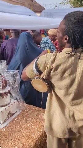 A video captures a joyous indoor event with a large gathering of people. In the foreground, a three-tiered white cake, beautifully adorned with dark brown frosting and intricate designs, sits on a sparkling gold tablecloth. The cake is wrapped in clear plastic, awaiting its reveal. To the right, a person in a tan traditional outfit, with dreadlocks, plays a small drum, contributing to the festive atmosphere. Their back is to the camera. The background is bustling with activity. Several women, dressed in colorful traditional African attire, are seen smiling and interacting. One woman in a white head wrap and a white lace dress is speaking into a microphone, her words likely guiding the celebration. A woman in a blue abaya stands nearby, while men in traditional Nigerian attire are also visible among the guests. The event space is decorated with white and clear awnings, suggesting an outdoor or semi-outdoor setup. The overall mood is cheerful and celebratory, indicating a wedding or a similar significant occasion.