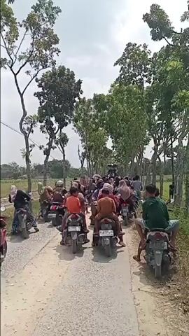 The video shows a group of people riding motorcycles on a dirt road. The people are dressed in casual clothing. The road is lined with trees on both sides. The sky is cloudy and the sun is shining. There is a traffic jam of motorcycles on the road. The motorcycles are moving slowly and some of the people are looking back.