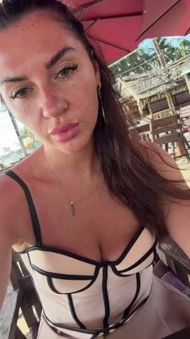 The video begins with a close-up selfie of a woman with long dark hair, wearing a light-colored corset-style top with black straps. She has freckles on her face and is wearing subtle makeup. She is sitting outdoors at a table, likely at a beachside establishment, as evidenced by the sand and beach umbrellas visible in the background. In the subsequent frames, the camera pans slightly, revealing more of the environment. There are wooden tables and chairs set up on the sand. Other people are visible in the background, including a man sitting at another table, wearing sunglasses and a patterned shirt. There are also some structures that appear to be part of a restaurant or bar. The woman is the primary subject throughout the video, and the background shifts as the camera moves.