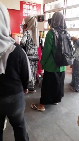 The video shows a group of people, mostly women wearing hijabs, standing in what appears to be a food establishment. The individuals are facing a counter where a person in a black shirt is working. There are menu boards with images of food and drinks displayed on the walls. The environment is well-lit with fluorescent lighting. The people are dressed casually, with some carrying backpacks. One child in a yellow outfit is also present. The scene suggests a queue or waiting area for service.