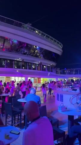 The video captures a lively night scene on what appears to be a cruise ship deck.  Multiple levels of the ship are visible, with the upper decks featuring illuminated railings and windows reflecting colorful lights.  The main area shown is an outdoor deck space at night, illuminated by a mix of purple and pink lighting, suggesting a party or event atmosphere.  A crowd of people are present, with many actively dancing in the center of the deck.  Several individuals are seated at tables with drinks, observing the activity or engaged in conversation.  A woman in a bikini is walking across the dance floor.  The dark night sky serves as the backdrop, with a visible wire extending across the top of the frame.