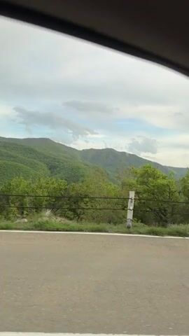 A car drives on a road bordered by a roadside fence with green mountains in the distance, beneath a cloudy sky. Then the camera focuses on a brown-haired, bearded man wearing a black t-shirt in the driver's seat, with the seatbelt buckled. He seems to be driving.
