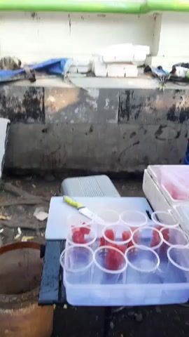 A man in an orange and purple shirt is preparing drinks in plastic cups, placing slices of red fruit into them. There are several white styrofoam containers stacked behind him. Another person is seated in the background, looking at a phone. The scene appears to be an outdoor stall with a blue canopy.