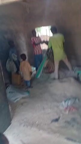 The video begins by showing the interior of a small, rough-hewn building. The walls appear to be made of mud or clay, and the floor is covered in dirt. Several people are inside, including two adults and two children. One adult, wearing a pink and white striped shirt, is interacting with a child in a yellow shirt. Another adult, in a lime green shirt, stands near a doorway. Various objects are visible, including sacks, a wooden box, and a green tarp or mat. The scene suggests a simple dwelling or storage area.

The video then transitions to an outdoor scene, focusing on several large sacks stored under a makeshift roof supported by wooden posts. The sacks appear to be agricultural products, with one clearly labeled "SUPER SALT" and "BAGCO". The surrounding area is rural, with trees and vegetation visible in the background, suggesting a village or farming community.

The camera then moves to show a sandy or dirt road with scattered debris. More trees and buildings are visible in the distance, indicating a settled area. The final shot shows a dusty clearing with several trees. The overall impression is of a rural Nigerian setting. The lighting suggests daytime, and the weather appears to be fair. The activity is not clearly defined but seems to involve people in a rural environment, potentially related to agriculture or trade given the sacks of goods.