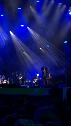 A live band performs on a stage bathed in blue lights. A female singer, dressed in a dark, ornate gown, stands center stage, holding a microphone. She has dark, curly hair and wears a large dark headdress. To her left, a male guitarist plays an electric guitar. Further to the left, a musician plays what appears to be a keyboard or synthesizer. Behind the singer and guitarist, two musicians stand with what seem to be string instruments, possibly violins or cellos. To the right of the singer, another musician is partially visible. The stage is elevated, with a dark backdrop. In front of the stage, a crowd of people is visible, their faces mostly obscured in shadow. The overall atmosphere is dark, with bright spotlights creating beams of light across the stage and through the haze.