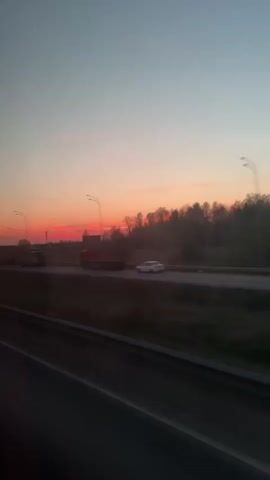 The video, taken from a moving vehicle, shows a highway at sunset. The sky is a gradient of pink, red, and pale blue. On the highway, several trucks and at least one white car are visible. There are streetlights along the highway, and a power transmission tower is also seen. A treeline is present on the horizon. The camera is positioned inside the vehicle, looking out the window.