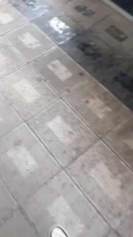 The video appears to be a first-person perspective of someone descending a set of wet outdoor stairs. The steps are made of stone or tile with darker borders. There are several vehicles visible at the top of the stairs, including a white van and a dark SUV. Behind the vehicles, a building is visible with the text "IXLOS MEDICAL CENTER" clearly displayed on its facade. The weather appears to be rainy, as evidenced by the wet surfaces and what look like raindrops falling. The time of day seems to be daytime, possibly overcast due to the rain and the lighting conditions. The scene is likely an urban or semi-urban environment, given the building and vehicles. No people are clearly visible in the video. The overall mood is functional, documenting the descent down the stairs in potentially inclement weather.