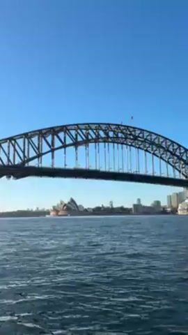 The video shows a view of the Sydney Harbour Bridge and the Sydney Opera House from the water on a clear, sunny day. The bridge is a large, steel arch structure spanning the harbor, and the opera house is visible in the distance with its distinctive white shell-shaped roofs. The water is relatively calm, with gentle ripples. The sky is a bright, clear blue. There's also a city skyline behind the opera house. The perspective suggests the video was taken from a boat or vessel on the water.