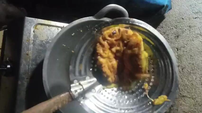A close-up overhead shot shows a person holding a perforated metal strainer with a wooden handle. Inside the strainer is a golden-brown, fried food item, possibly a fritter or omelet, that is roughly circular in shape. The food item is resting in a large, dark-colored frying pan, which is on a stovetop. The background is dimly lit and appears to be an outdoor or rustic kitchen setting, with a concrete-like surface visible to the right and a metal stove to the left. The video shows slight movements and shifts in perspective as the strainer is maneuvered.