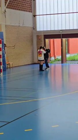 The video shows a man and a woman in a gymnasium. They are boxing. The woman is wearing a white t-shirt with graphics, black shorts, and black kickboxing boots. The man is wearing black sweatpants and a black t-shirt. He is holding boxing mitts. The gymnasium has a blue floor with yellow lines, and a light-colored brick wall is to the left. There is a wooden chair next to a blue and red pole. There is a large window and doors on the right. The environment outside appears to be green with a building that is orange.