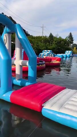A wide shot shows a floating inflatable water park on a lake. In the foreground is a blue and white inflatable slide with a red platform. Beyond that, several other inflatable structures are visible, including slides, obstacle courses, and a large red inflatable island. The lake is calm, with reflections of the sky and surrounding trees. In the background, lush green trees and rolling hills can be seen under a partly cloudy sky. The water park appears to be empty of people.