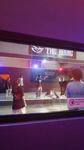 A group of five young people are practicing a dance routine on a stage. The stage is lit with purple and blue lights. There is a banner that says "THE KLAN" and an eye logo at the top of the stage. There is also a sign that says "2023 1ER LUGAR GRUPOS SENIOR DALIMAH". A person in the foreground is filming the dancers. The dancers are wearing casual clothing and appear to be focused on their choreography.