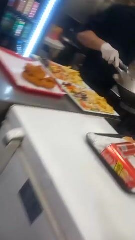 A cook wearing gloves is preparing plates of food in a kitchen. Two plates contain salad, chicken, and french fries. A third plate contains chicken tenders and french fries. The cook uses tongs to move french fries from a metal bowl to the three plates. There are canned drinks and hamburger buns on a tray in the background.